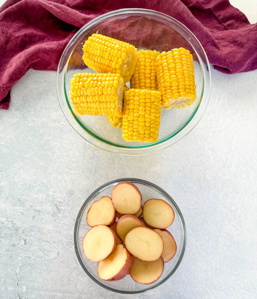 fresh corn on the cob and sliced red potatoes in separate glass bowls