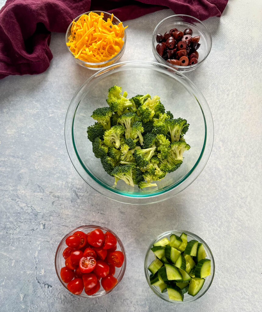 shredded cheddar cheese, sliced olives, broccoli, tomatoes, and diced cucumbers in separate glass bowls