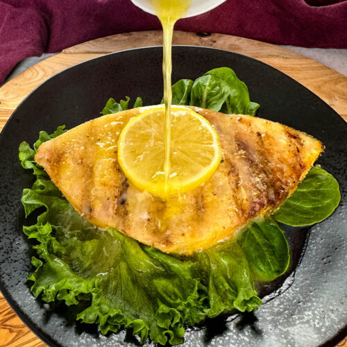 smoked swordfish with lemon and greens on a black plate drizzled with lemon butter