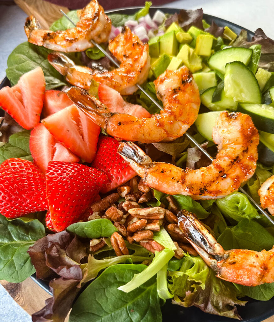 grilled shrimp salad on a plate with mixed greens, strawberries, avocado, and onions