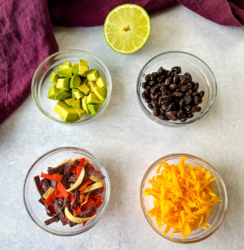 fresh lime, avocados, black beans, tortilla chips, and shredded cheese in separate glass bowls
