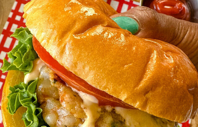 person holding shrimp burger on a brioche bun with lettuce, tomatoes, and spicy mayo