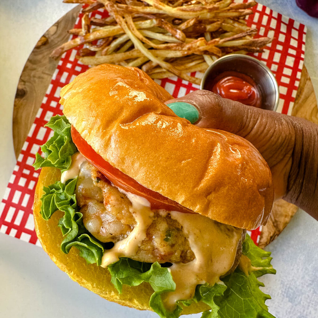 person holding shrimp burger on a brioche bun with lettuce, tomatoes, and spicy mayo