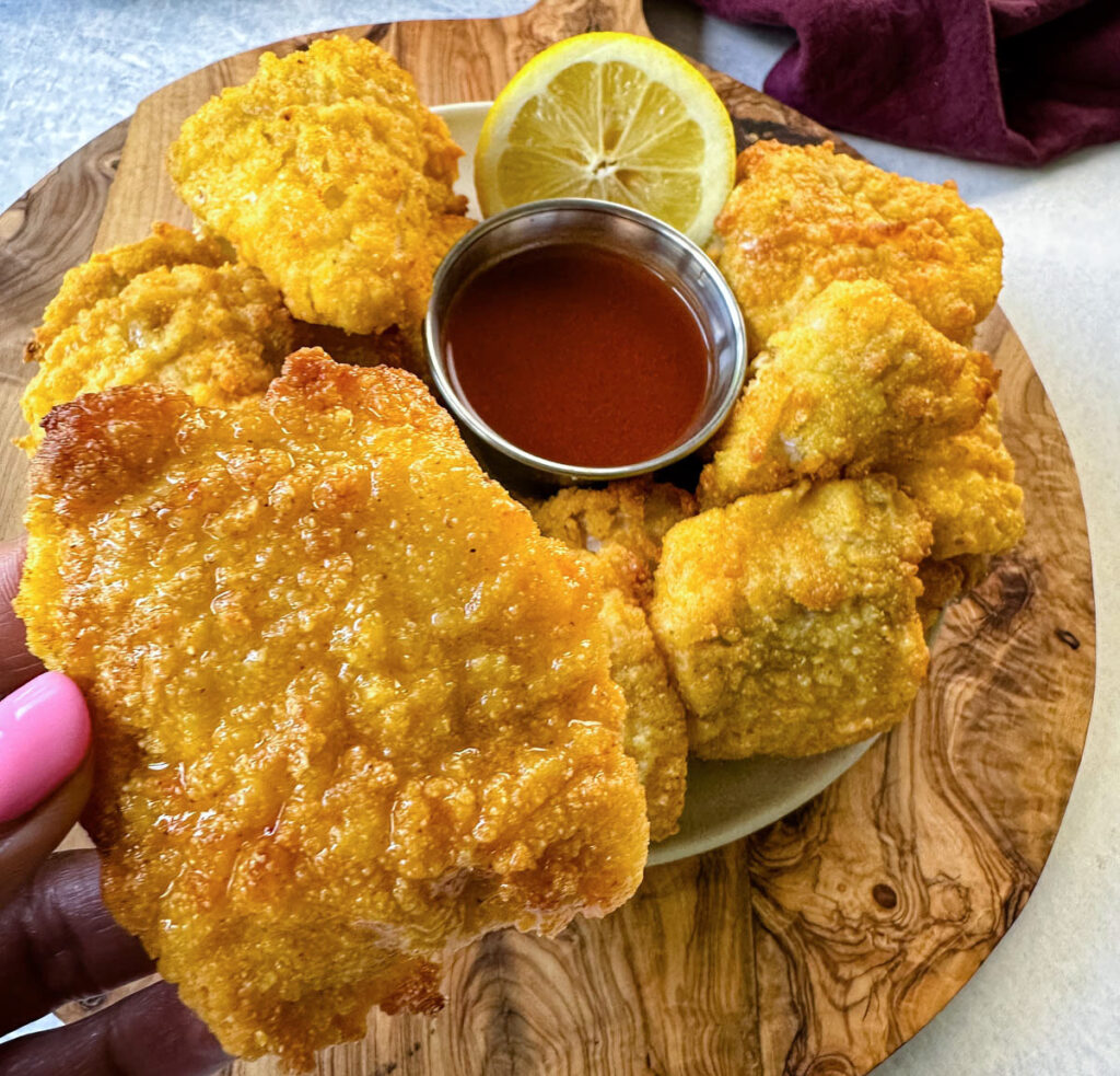 person holding breaded catfish nugget with hot sauce and a fresh lemon