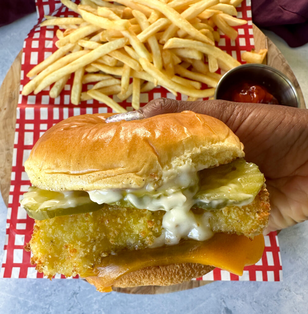 person holding fish sandwich with cheese, pickles, and tartar sauce on a brioche bun on a plate with fries