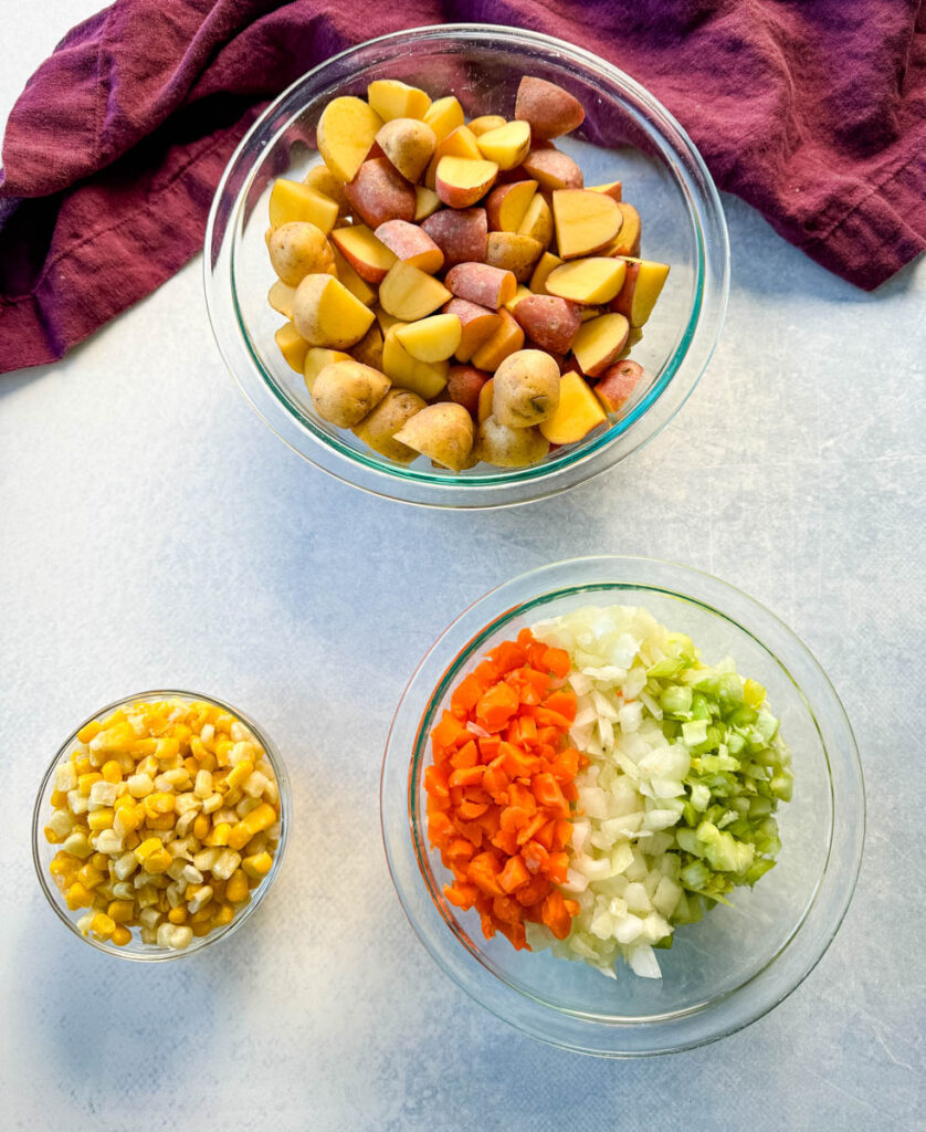 raw potatoes, corn, carrots, celery, and onions in separate glass bowls
