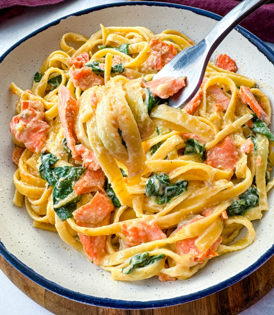 a forkful of salmon Fettuccine alfredo with spinach over a white bowl